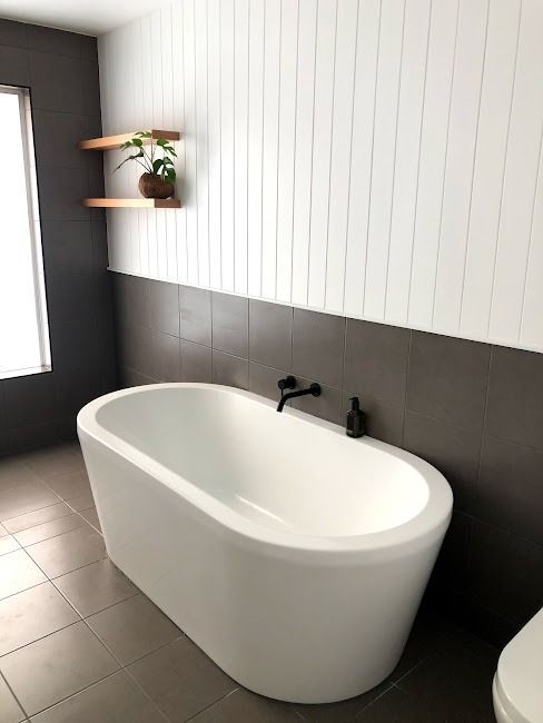 A modern bathroom featuring a white freestanding oval bathtub against a dark tiled wall and white vertical paneling.