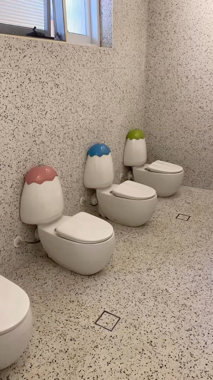A row of three small toilets with pink, blue, and green rounded lids in a bathroom with speckled walls and floors.