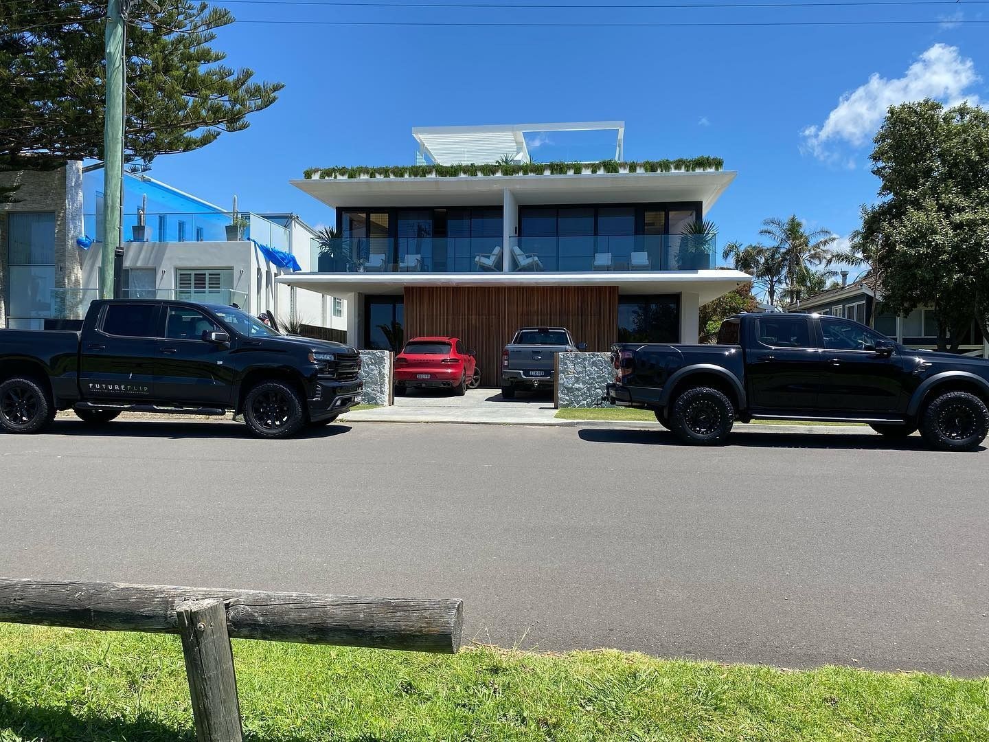 A two-story, modern home with a balcony and wooden garage doors, flanked by two black trucks parked on the street.