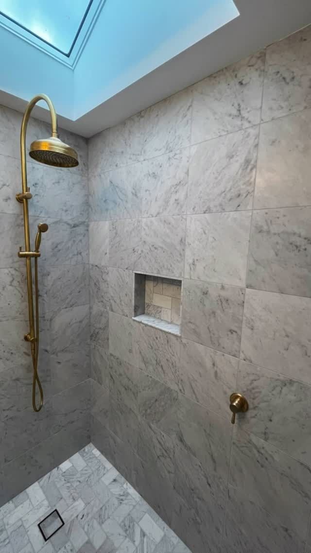 A shower interior with grey marble-tiled walls and floor, a brass showerhead, matching fixtures, and a recessed niche.