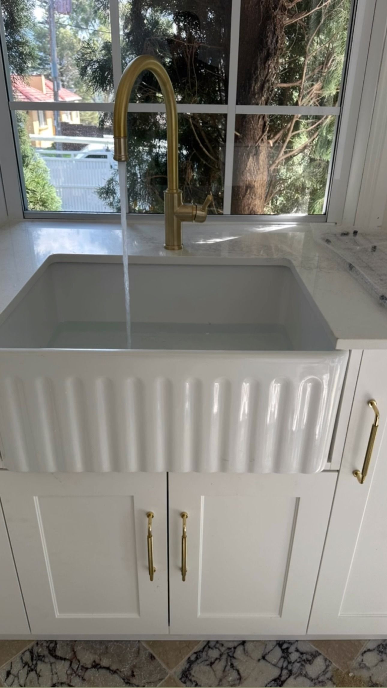 A white fluted farmhouse sink with a gold faucet running water, set in a kitchen with white cabinetry and a window.