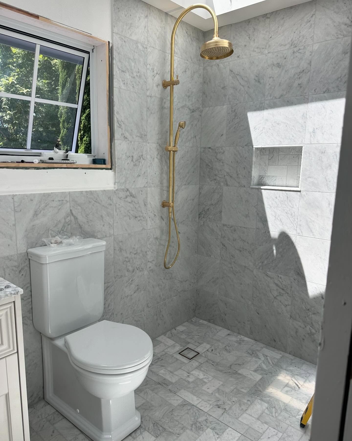 A bathroom with grey marble-patterned tiles, a white toilet, and a gold rainfall shower head inside a shower area.