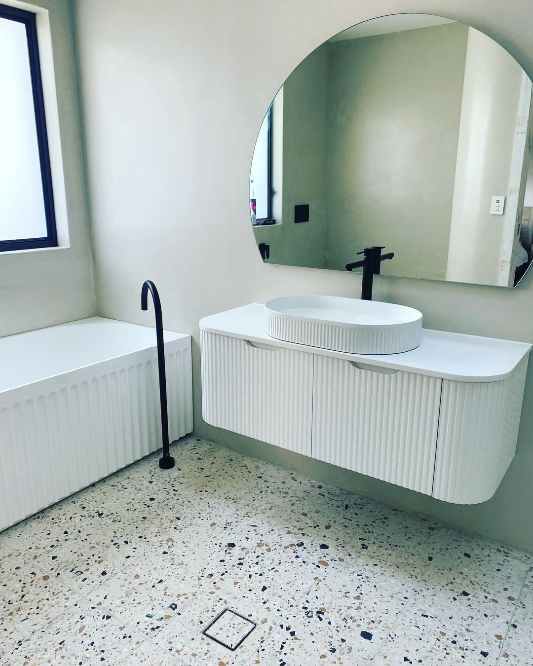 A modern bathroom with a fluted white floating vanity, an arched mirror, terrazzo flooring, and a freestanding bathtub.