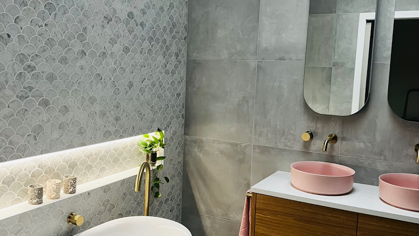 A bathroom featuring gray scalloped and slab wall tiles, a light-filled recessed shelf, and two pink vessel sinks.