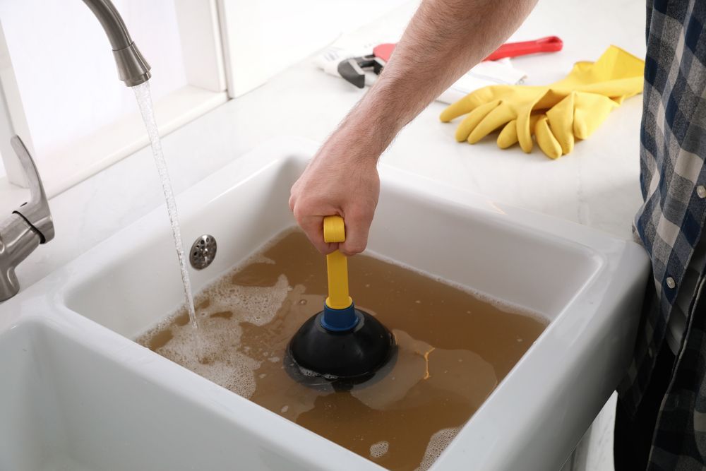 A person uses a plunger in a kitchen sink filled with murky water while a faucet runs.