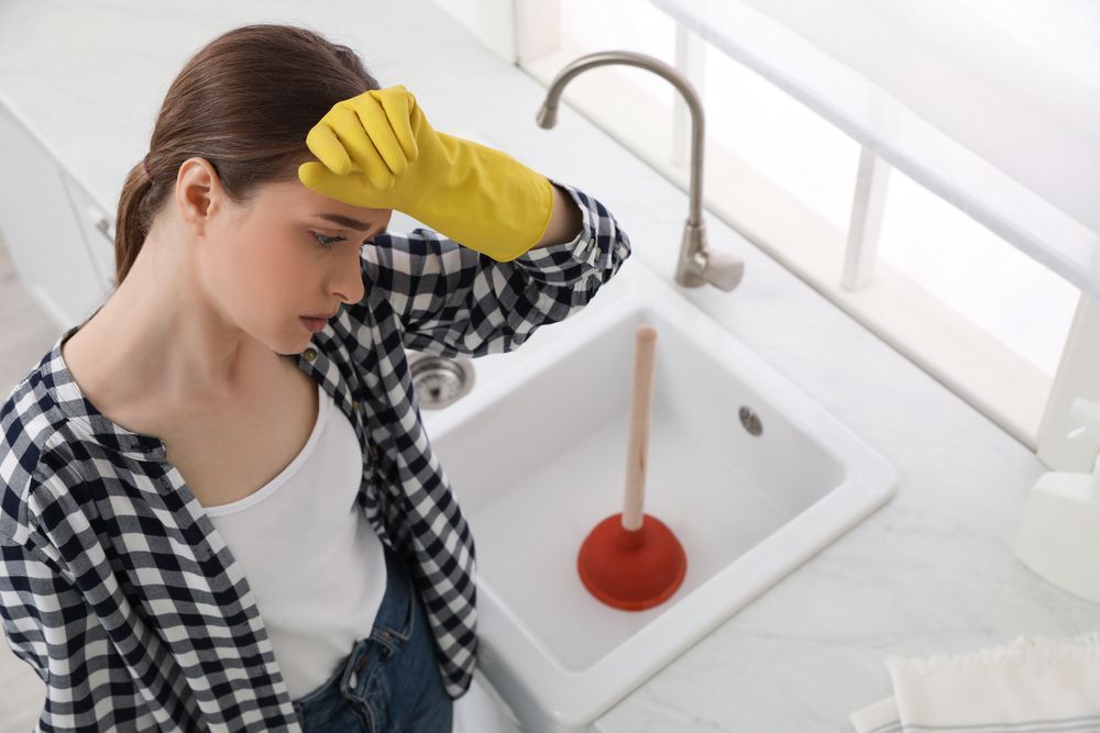 A person wearing a yellow rubber glove looks frustrated while holding a plunger in a white kitchen sink.