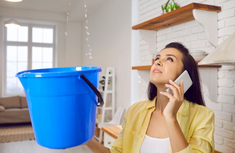 A person looks up at a ceiling leak while holding a blue bucket and talking on a smartphone in a kitchen.