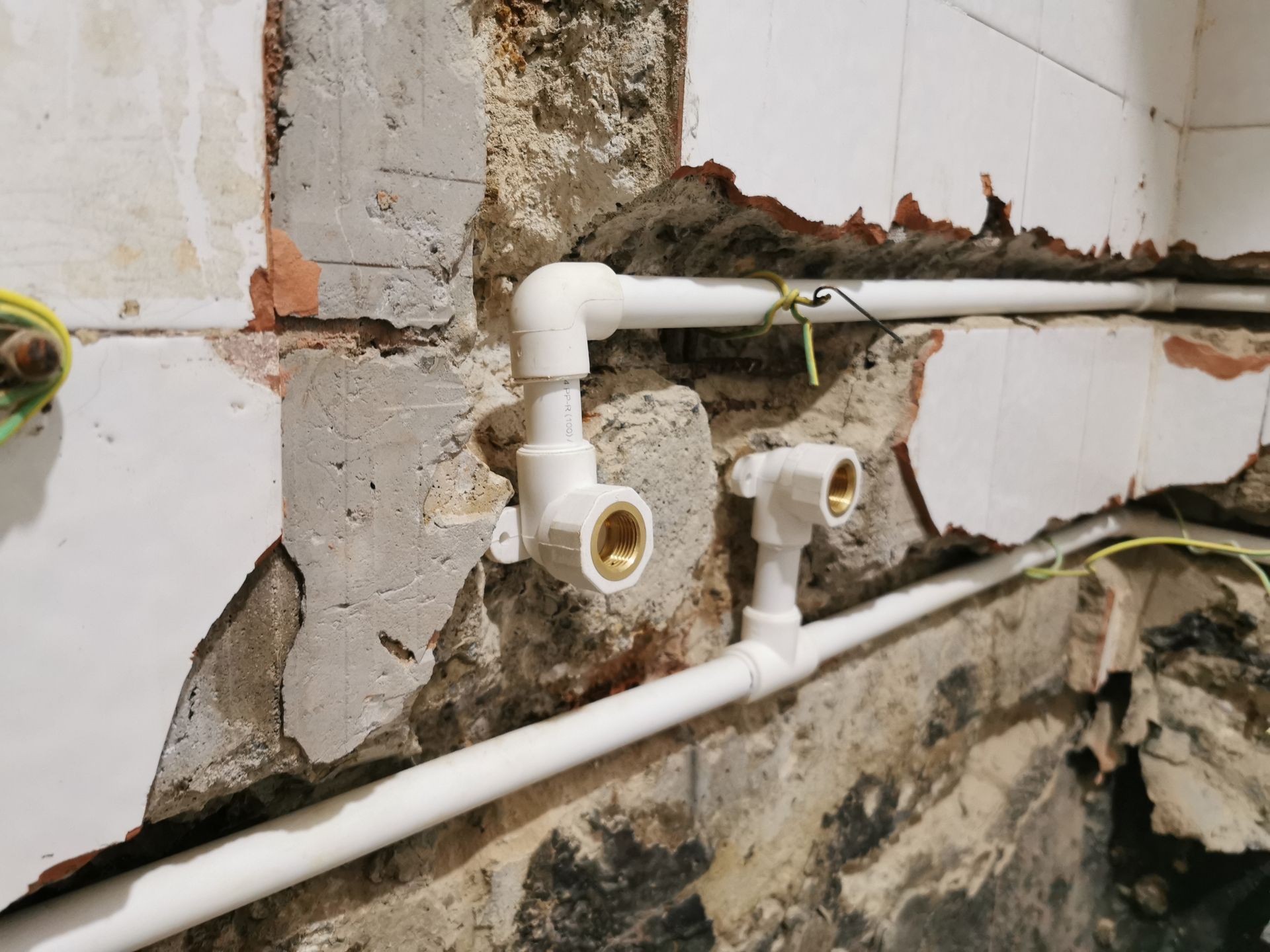 Exposed white PEX piping and brass-threaded fittings installed against a damaged, unfinished wall.
