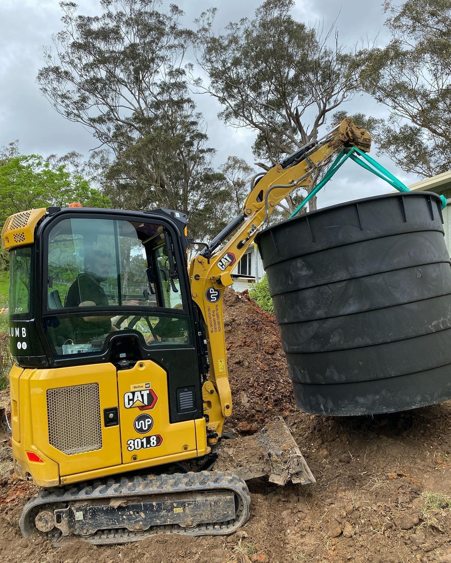 A yellow CAT mini excavator using green slings to lift a large, black plastic tank on a construction site.