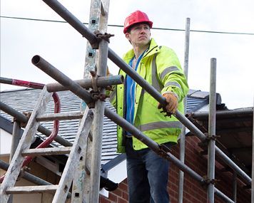 A man in a yellow jacket and red hard hat is standing on a scaffolding.