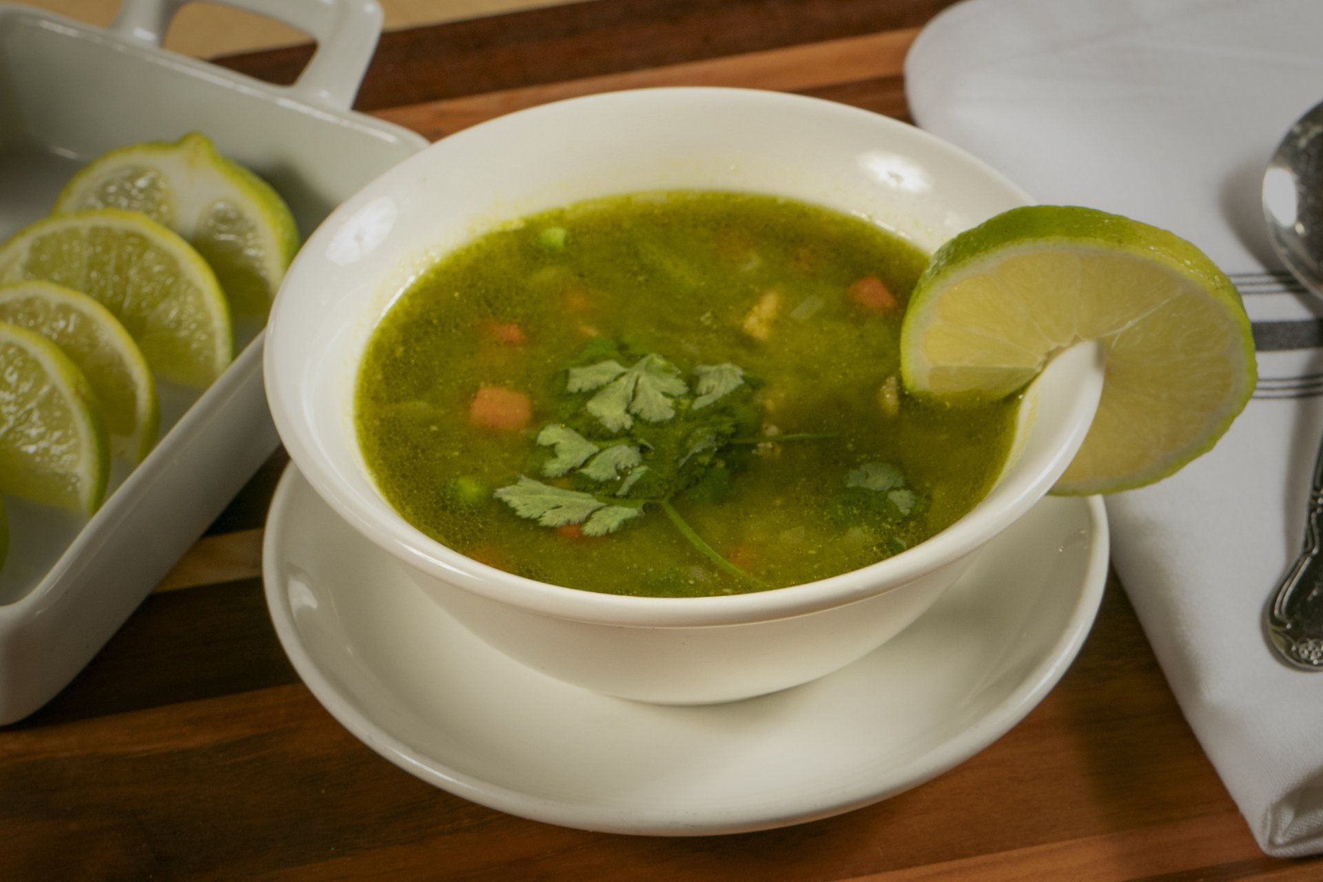 Bowl of green soup garnished with lime and cilantro, on a wooden surface with lemon slices.