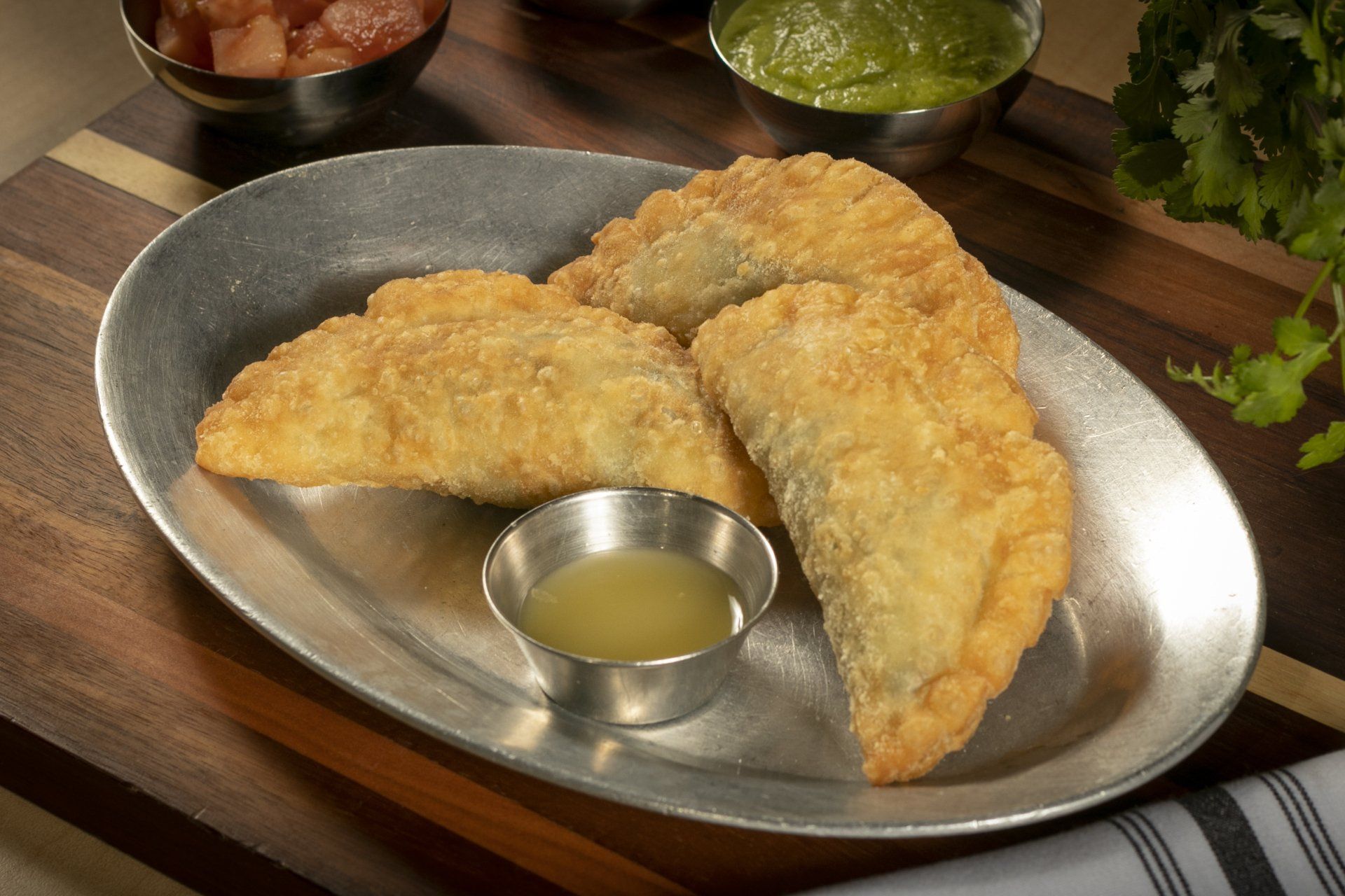 Three golden-brown empanadas on a metal platter with a small dipping sauce, salsa, and guacamole.