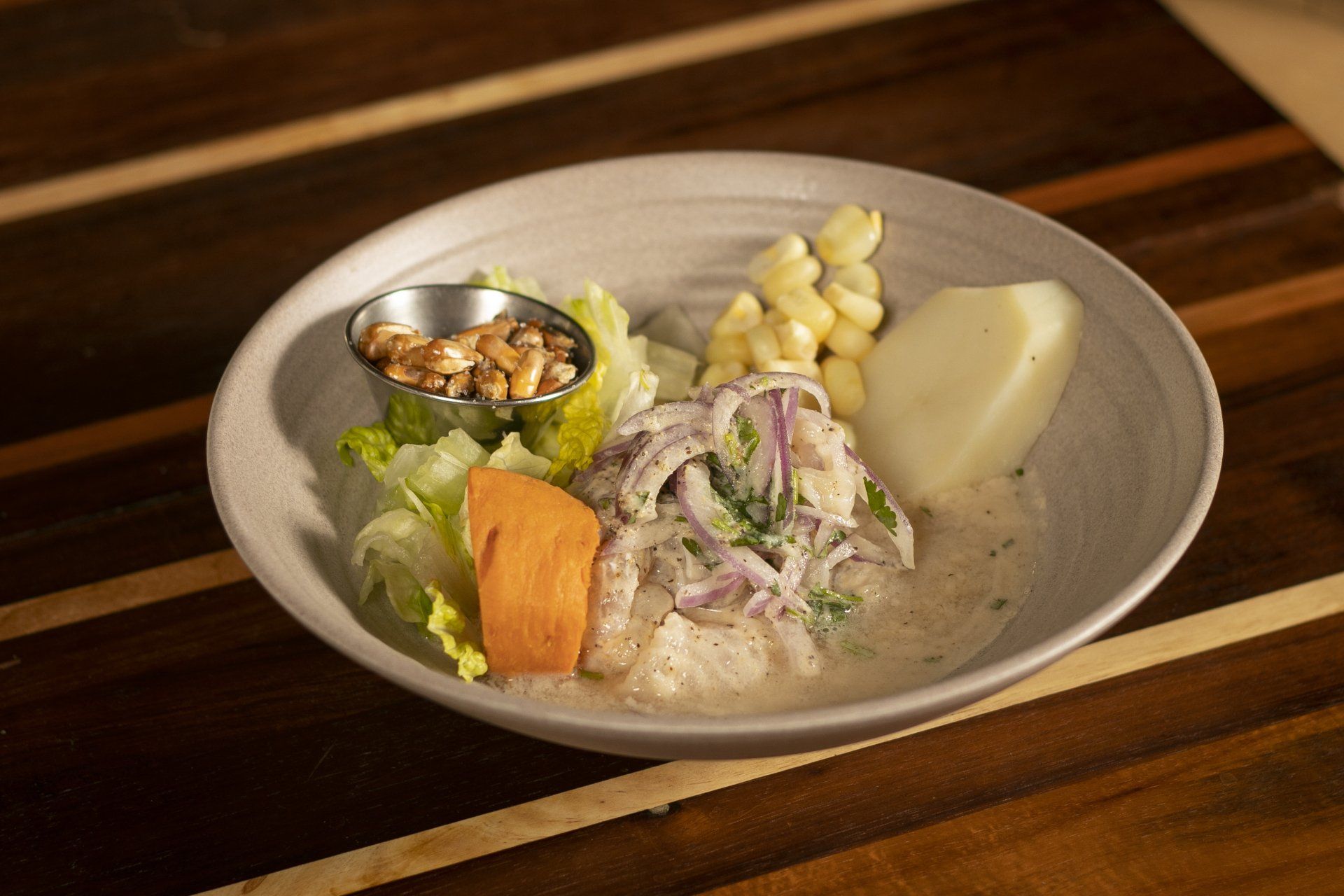 Ceviche dish with fish, corn, sweet potato, and garnish in a grey bowl on a wooden table.