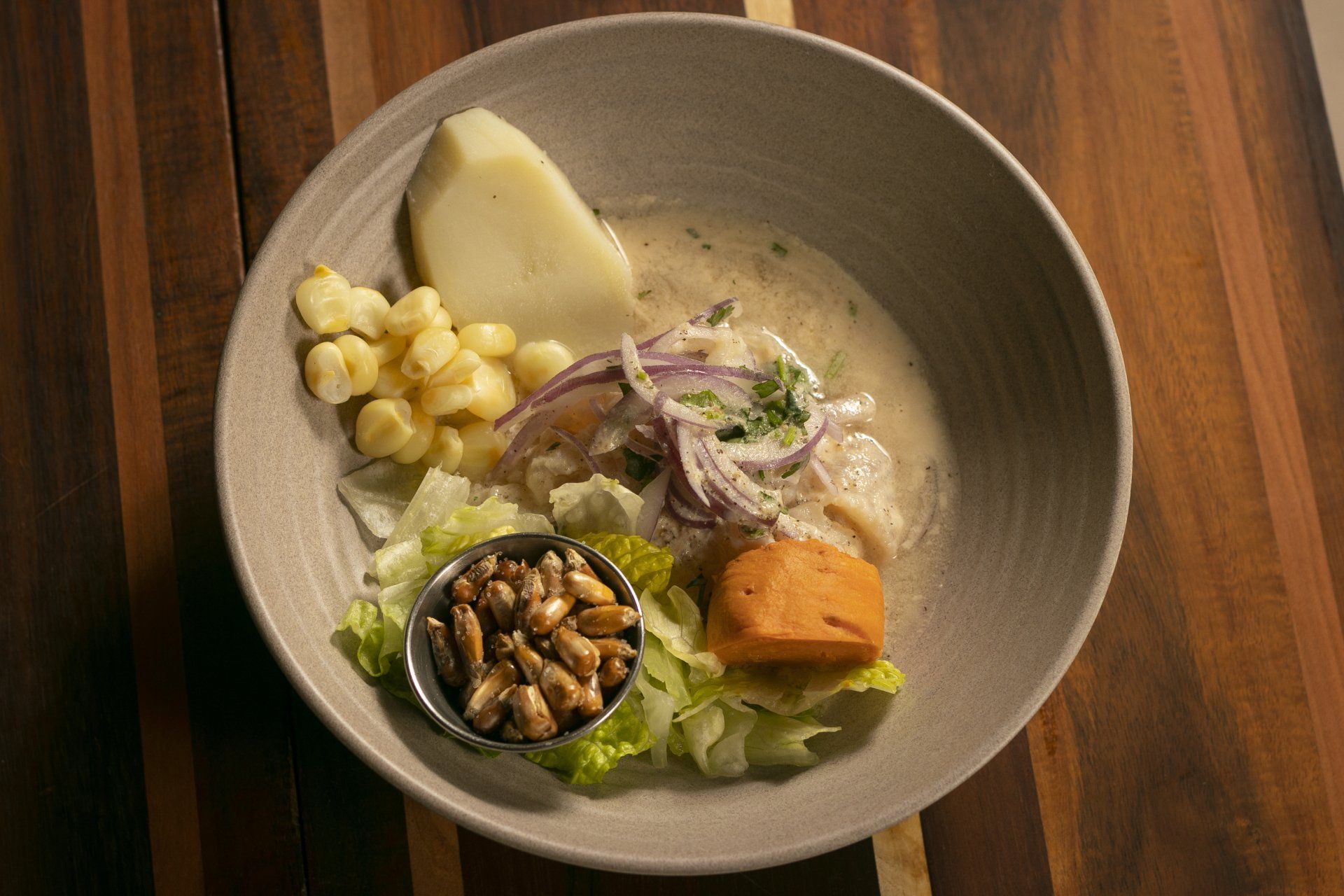 Ceviche served in a grey bowl, with purple onion, sweet potato, corn, and potato.