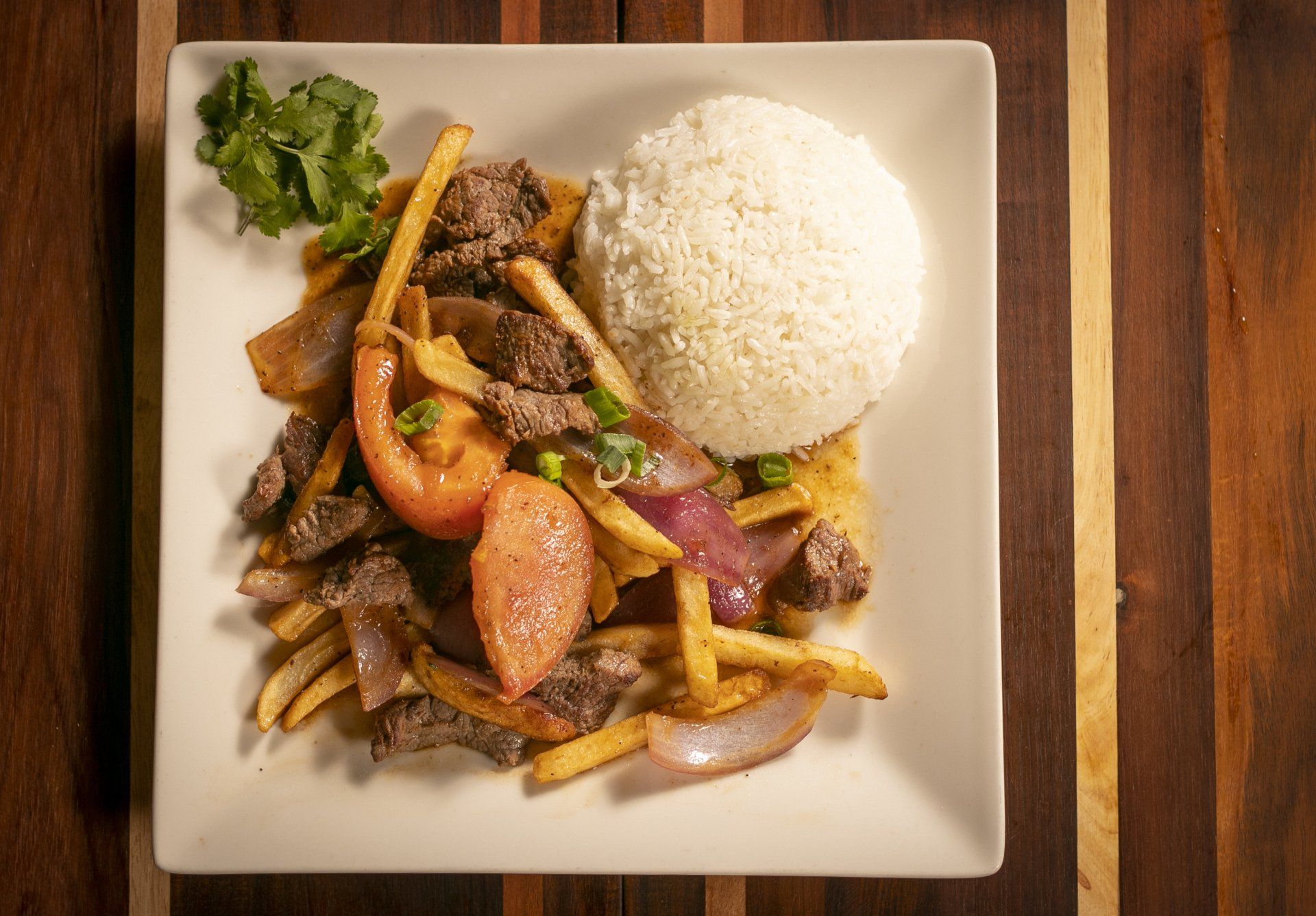 Lomo saltado with rice, steak, fries, red onion, and tomato on a square white plate, wooden background.