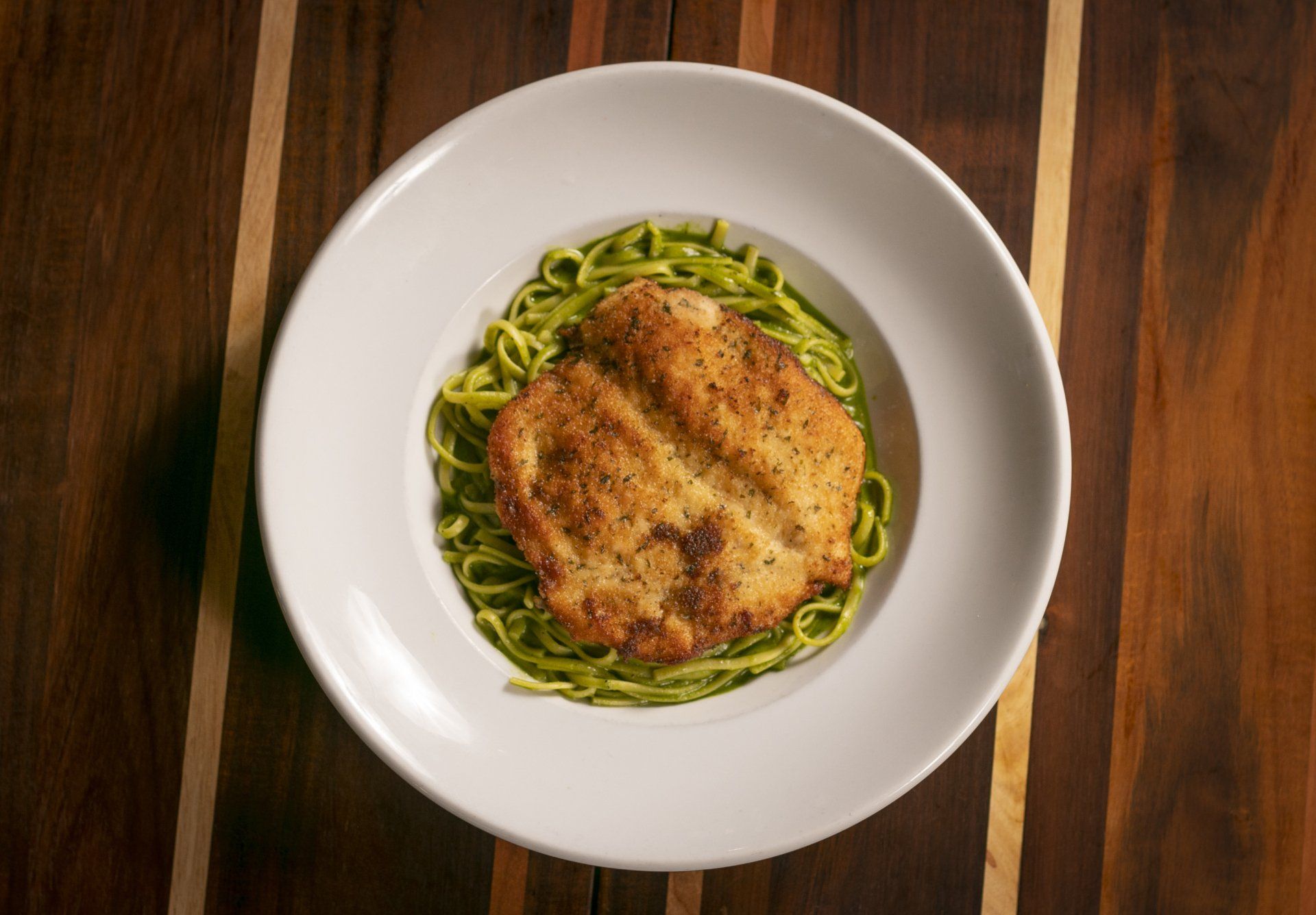 Chicken cutlet on green pasta in a white bowl, set on a wooden surface.