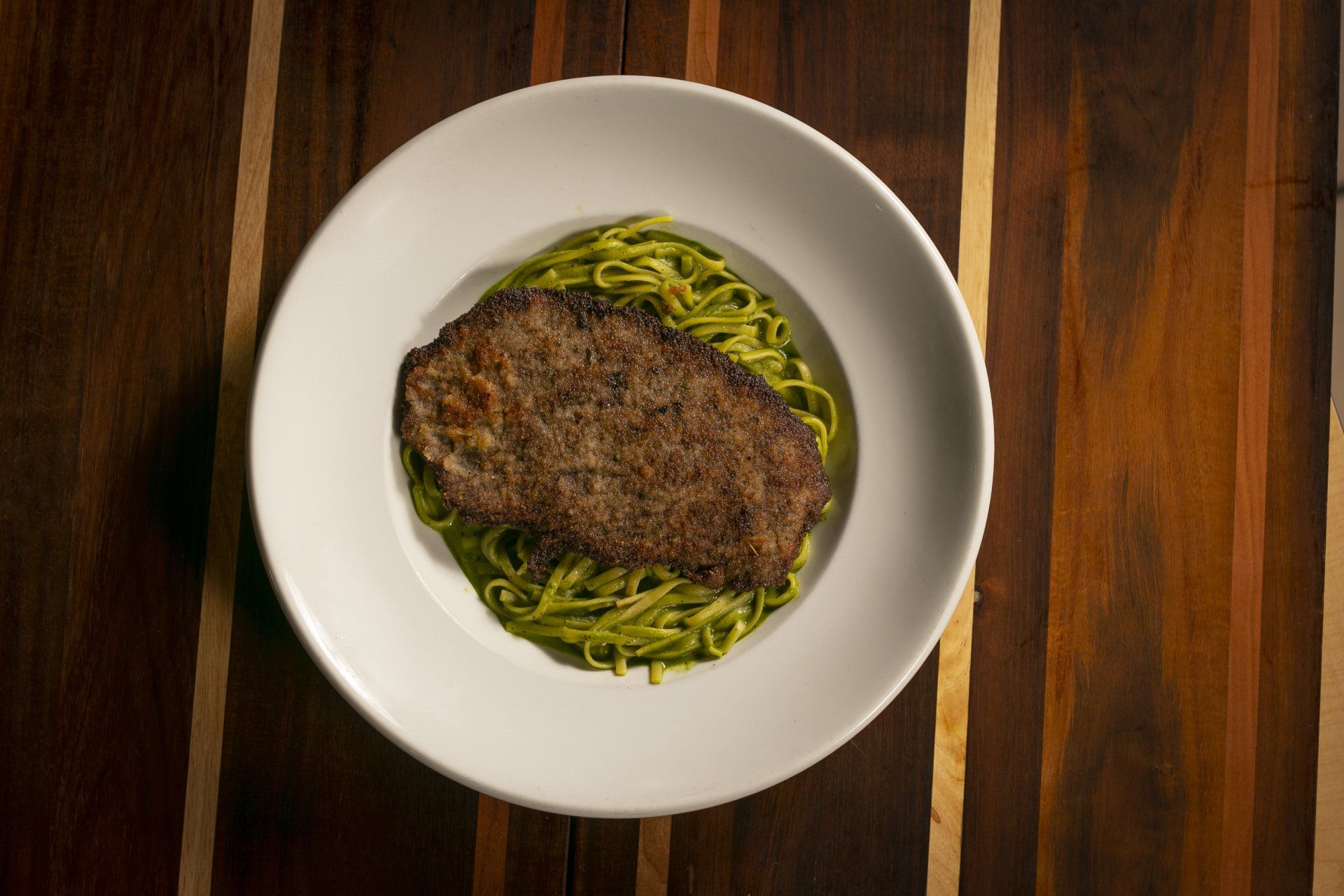 Steak on a bed of green pasta in a white bowl, on a wooden table.