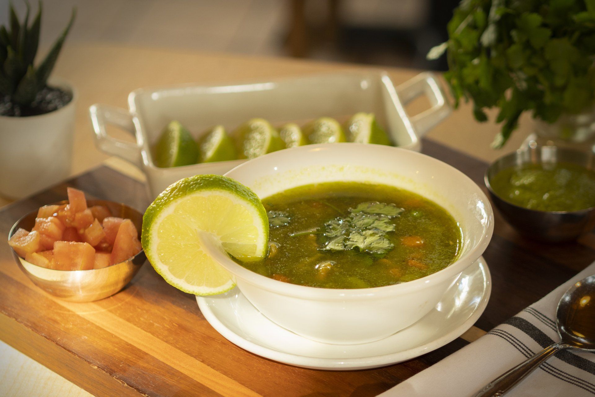 Bowl of green soup with lime wedge, cilantro, and diced tomatoes on a wooden board.