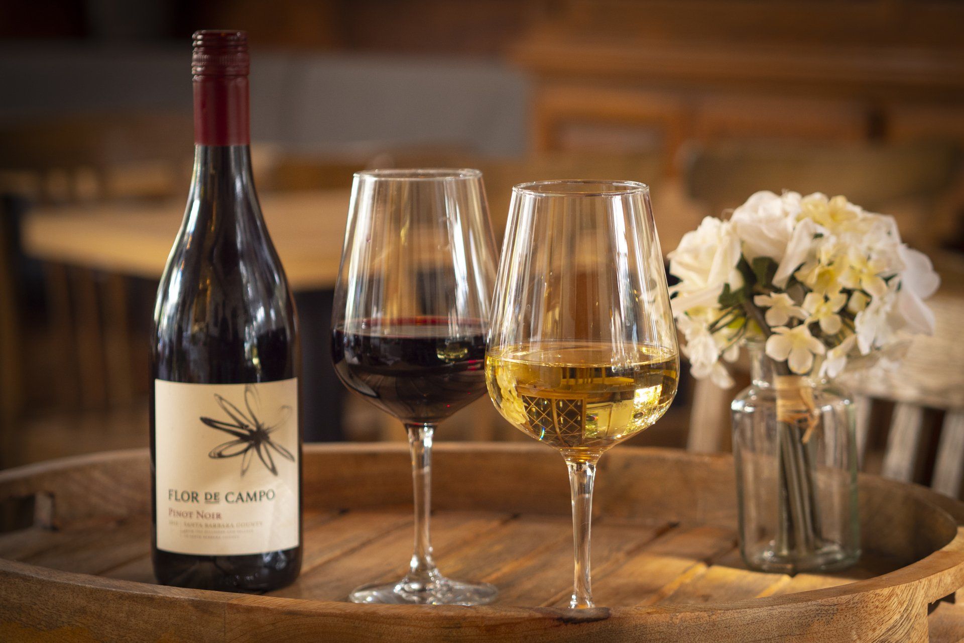 Wine bottle, two glasses of red and white wine, and flowers on a wooden tray.