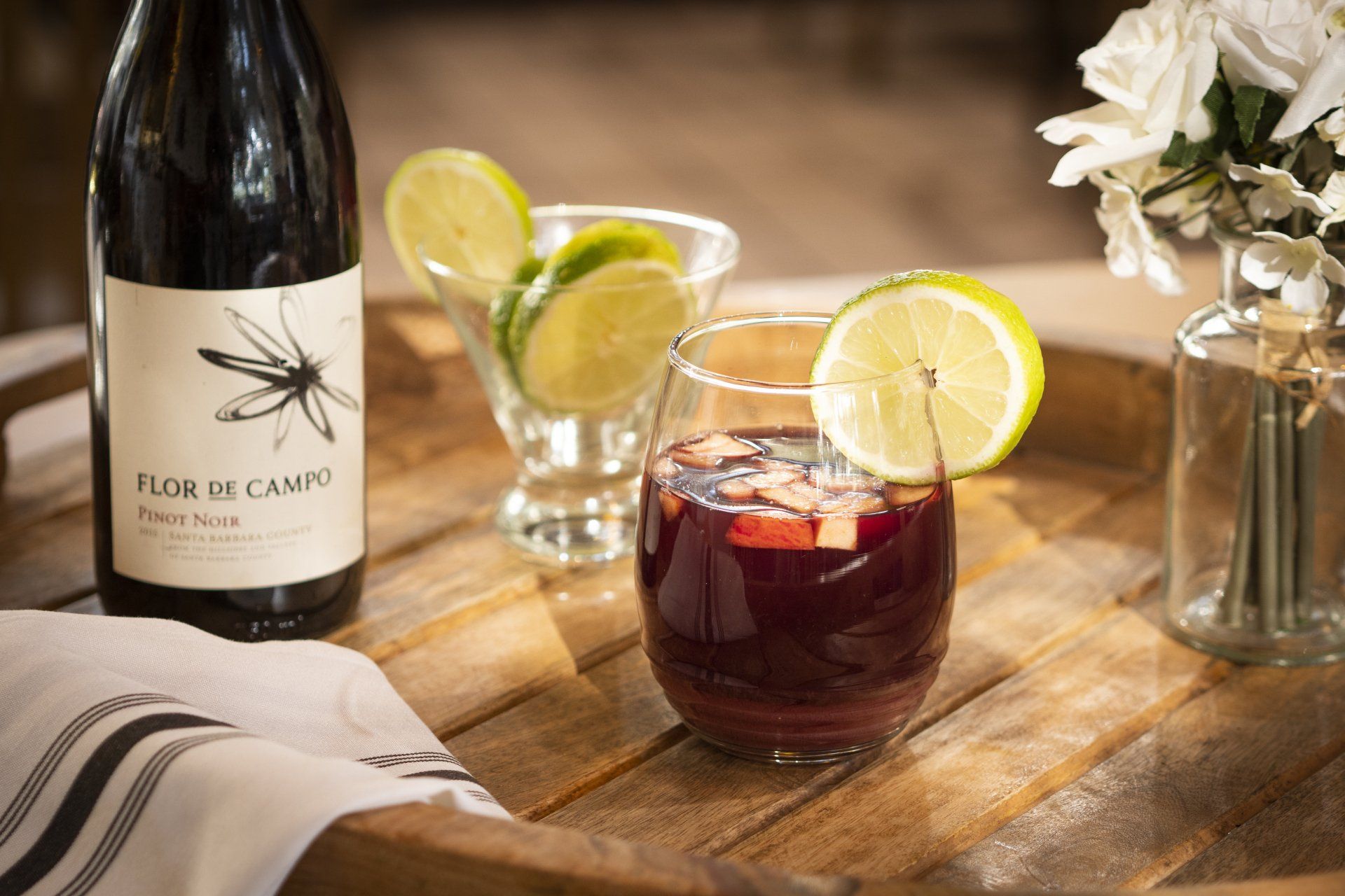 A glass of red sangria with a lime, next to a wine bottle and a vase of white flowers on a wooden tray.