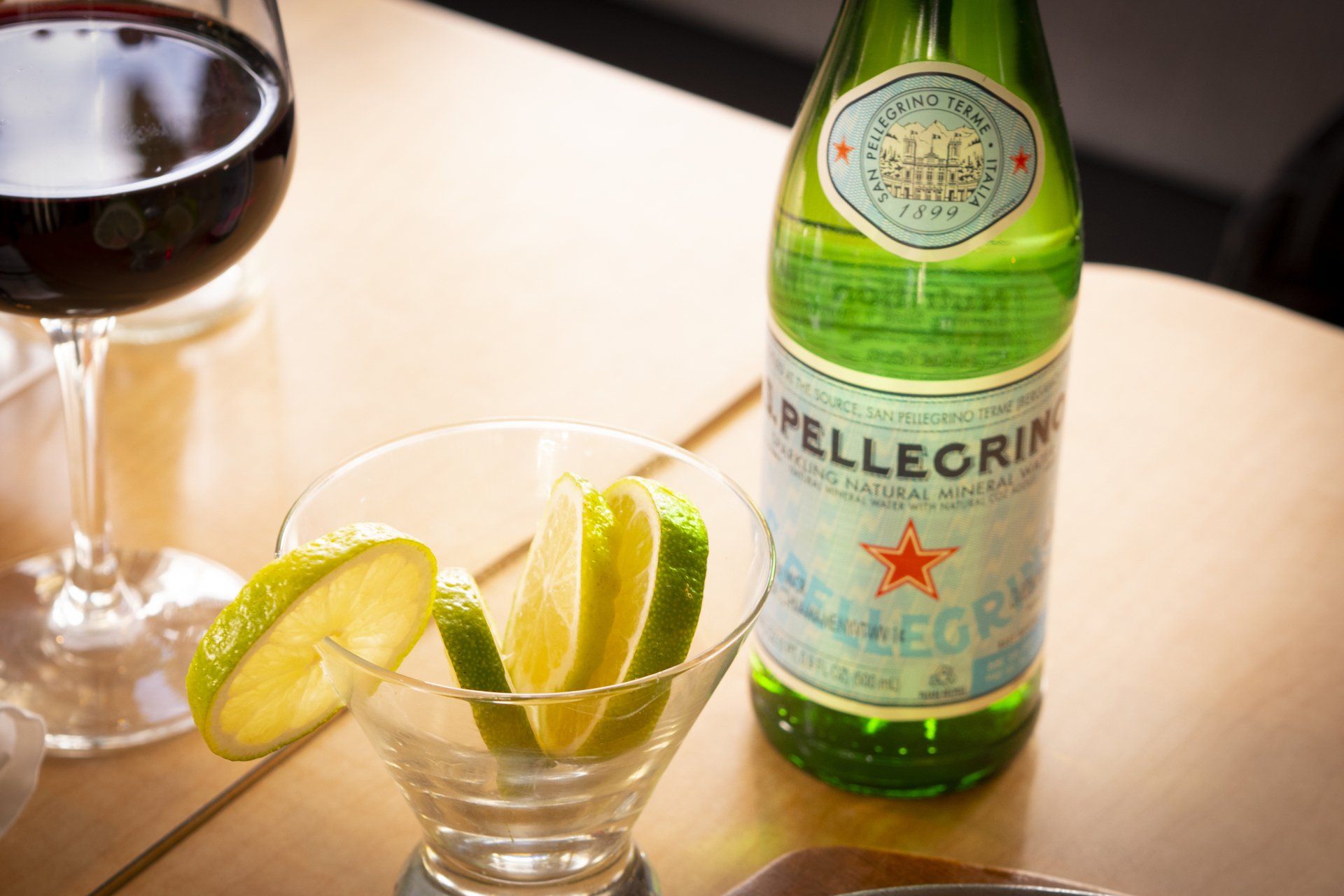 Bottle of San Pellegrino, lime slices in glass, red wine glass on a table.