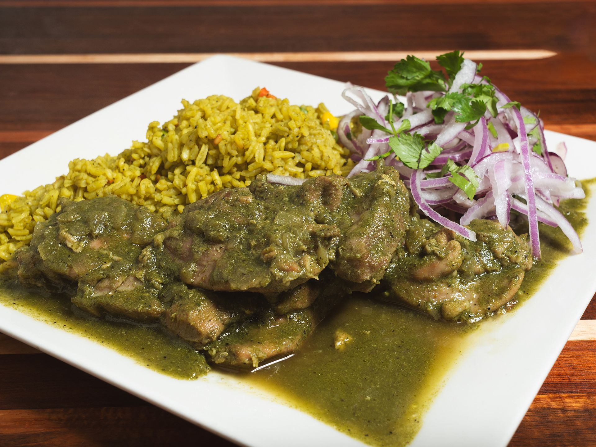 Sliced meat in green sauce, yellow rice, red onions, cilantro on a white plate.