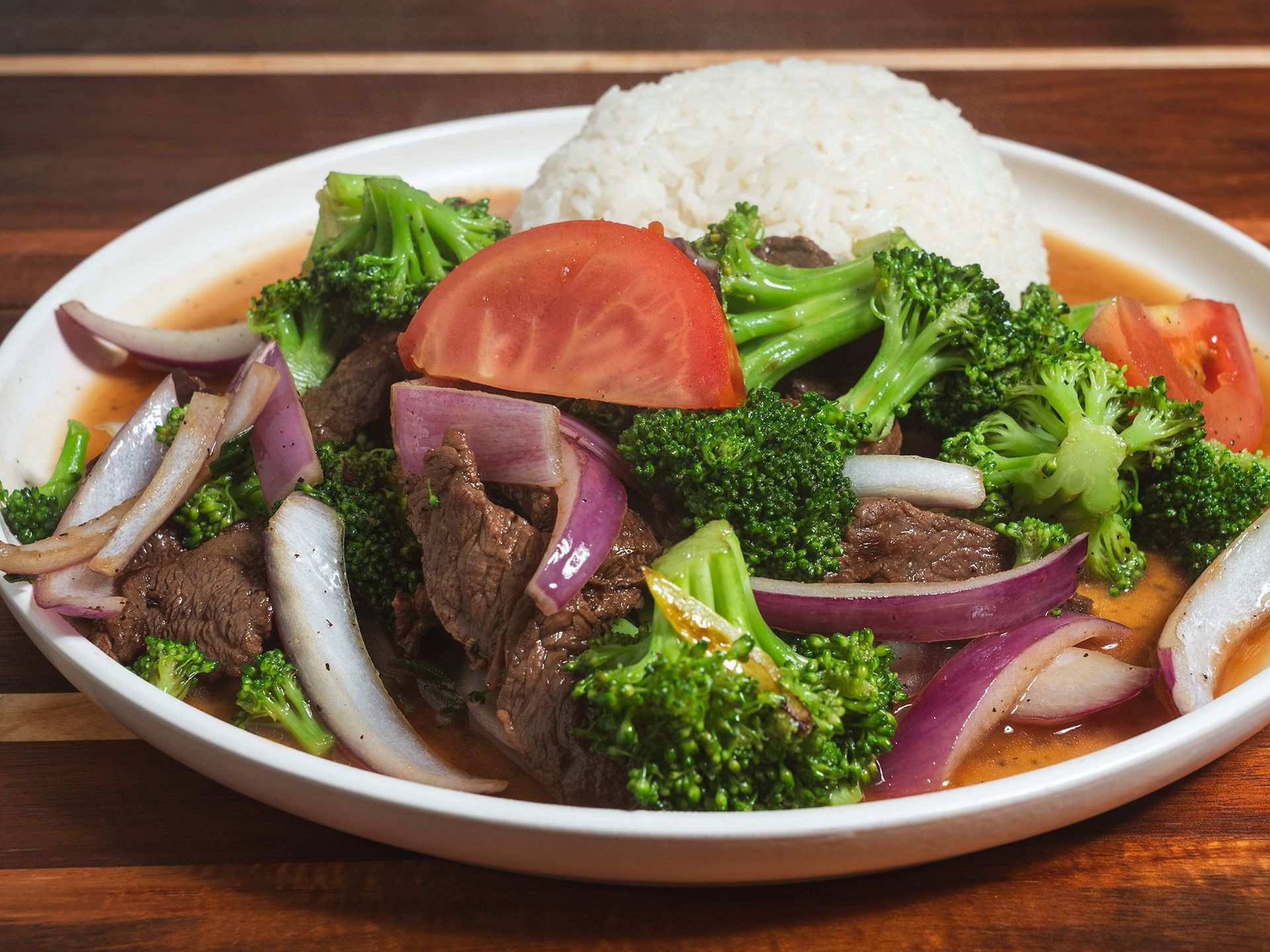 Beef and broccoli stir-fry with red onion, tomato, and rice on a white plate.