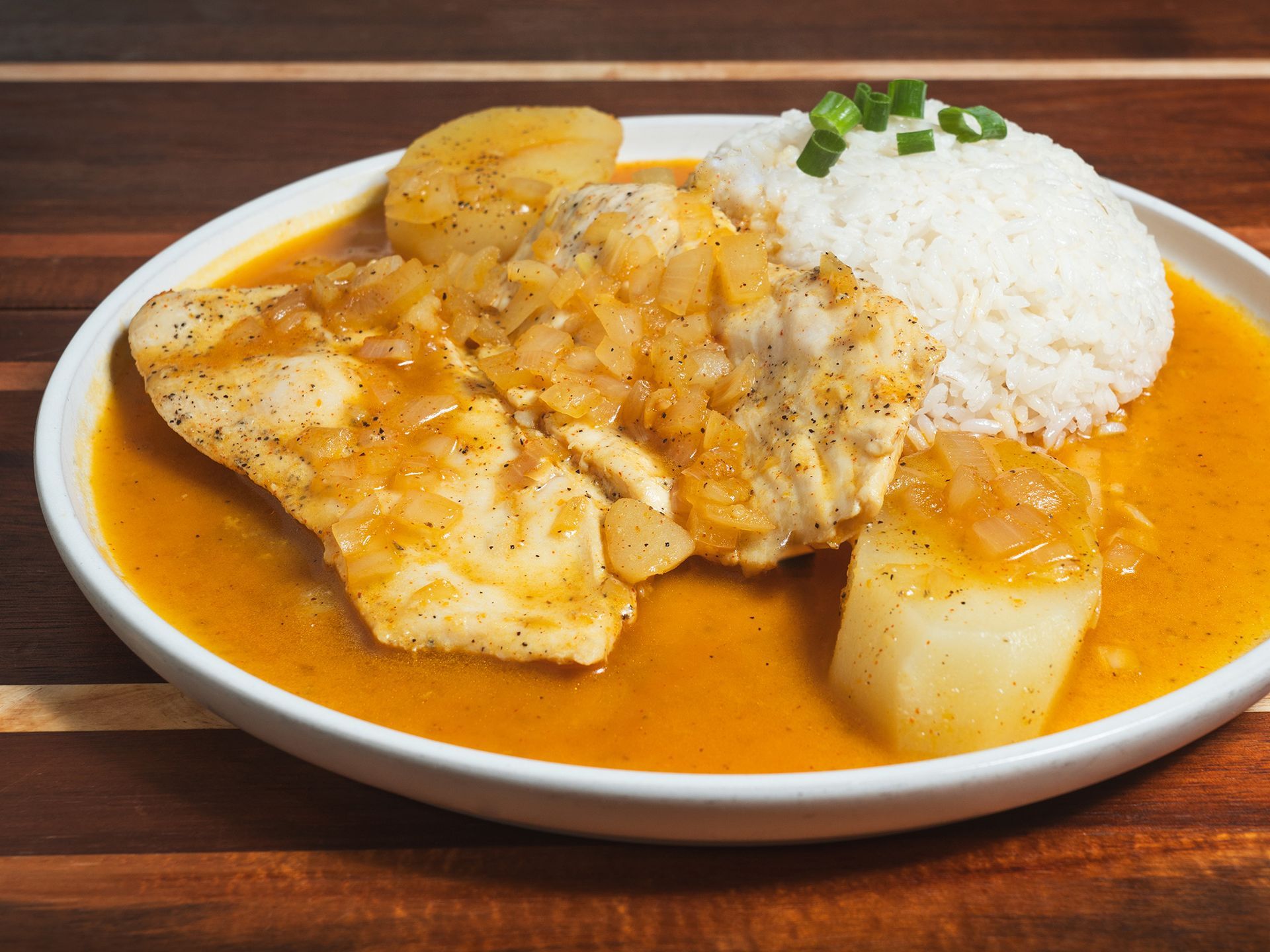 Fish in sauce with rice and potatoes, garnished with green onions, on a white plate.