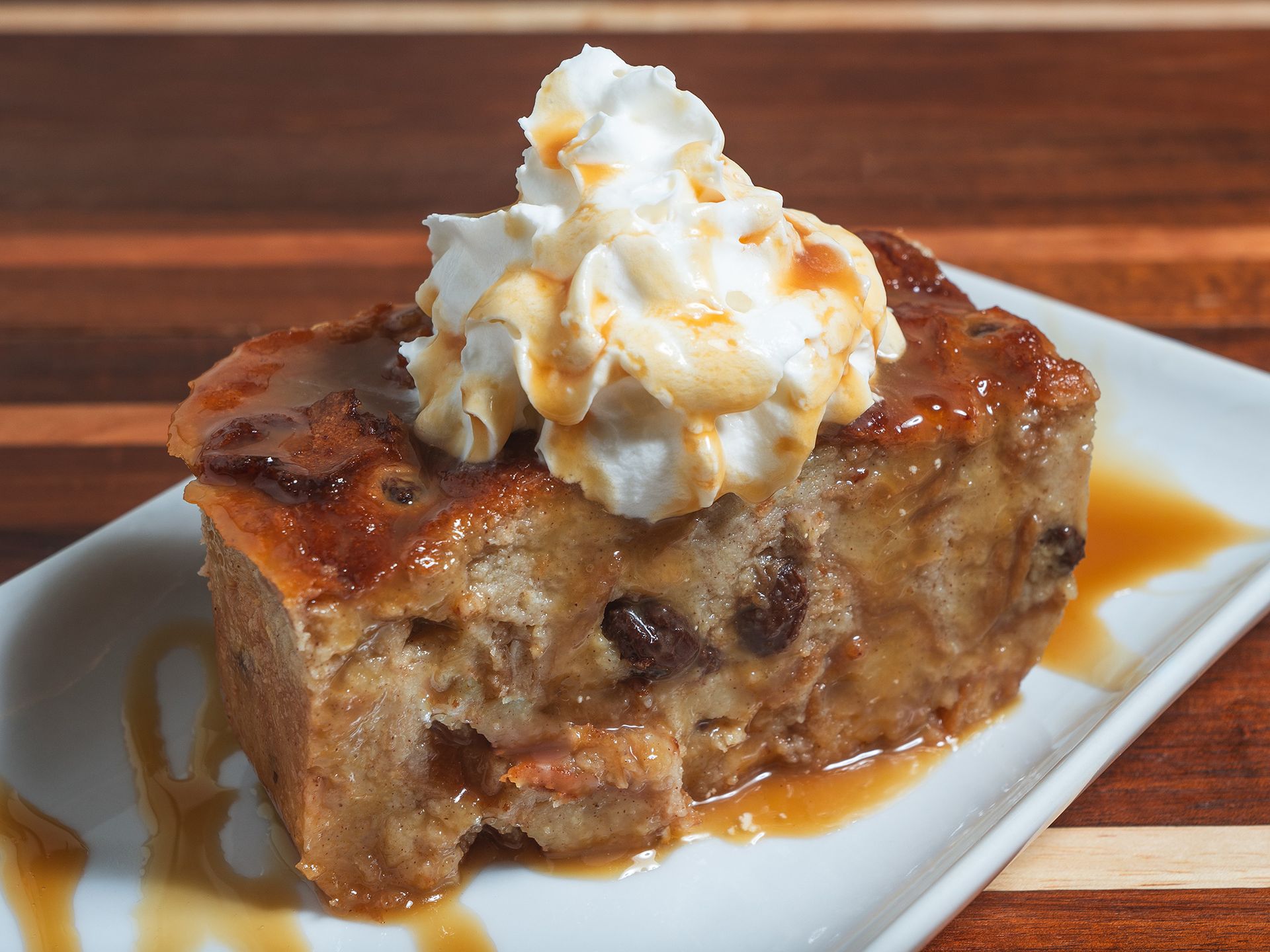 Bread pudding topped with whipped cream and caramel sauce on a white plate.