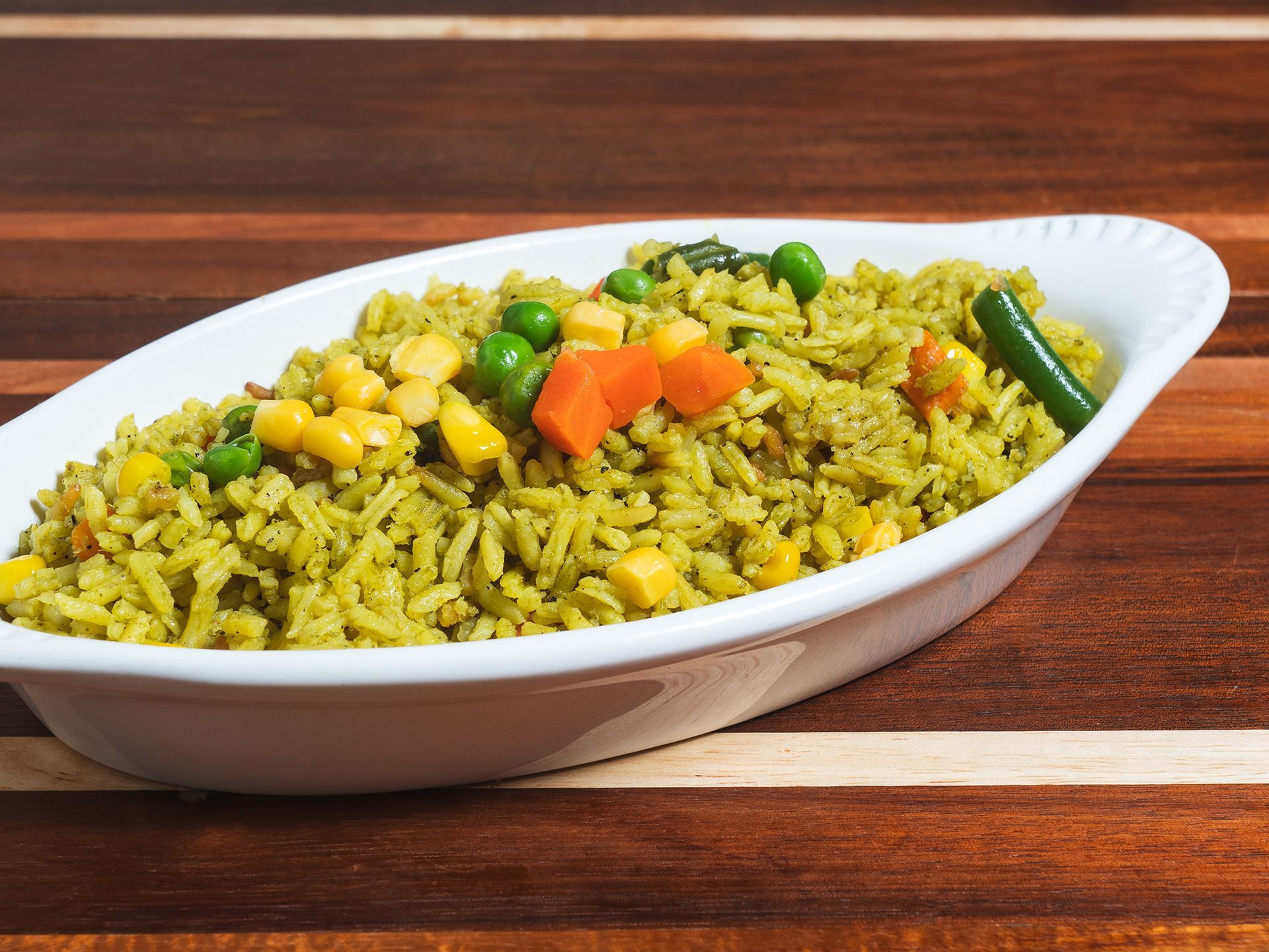 Green rice dish with peas, carrots, corn, and a green chili pepper in a white bowl on wood.