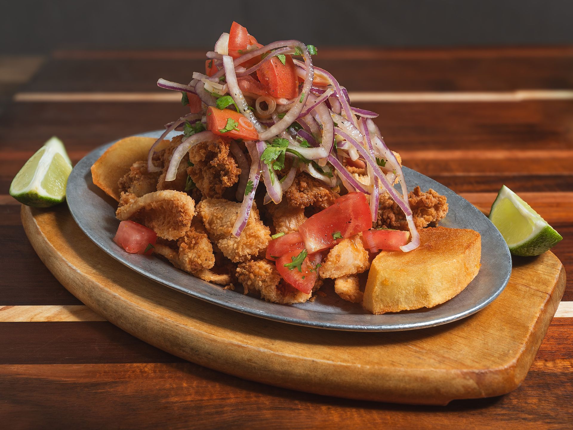 Fried seafood dish with tomato and onion topping, lime wedges, on a wooden board.