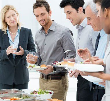 People at a buffet serving themselves food, indoors. Several men and one woman, some holding plates.