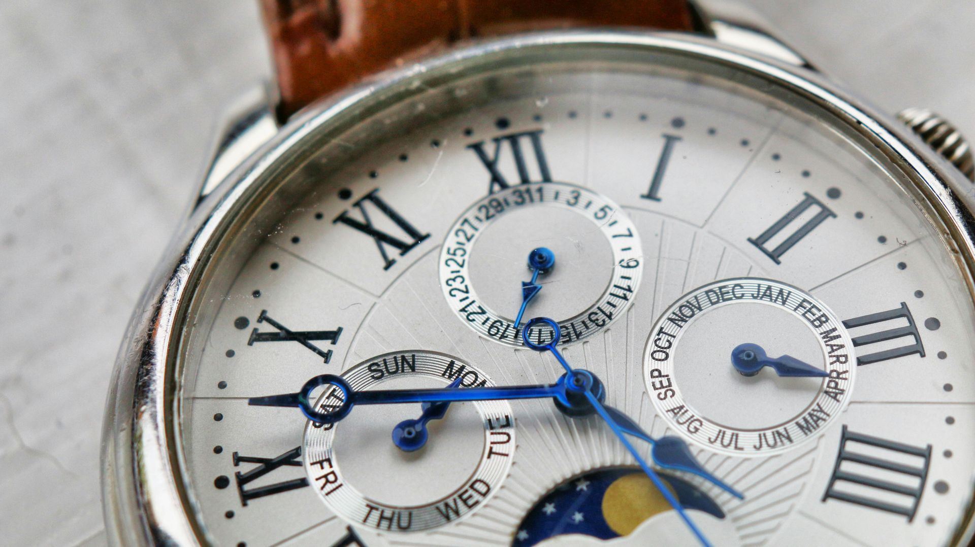 Close-up of a silver wristwatch with a white face, Roman numerals, and subdials. Blue hands and brown leather band.
