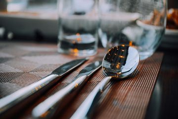 Close-up of silverware, including a spoon and two knives, on a brown placemat with water glasses in the background.
