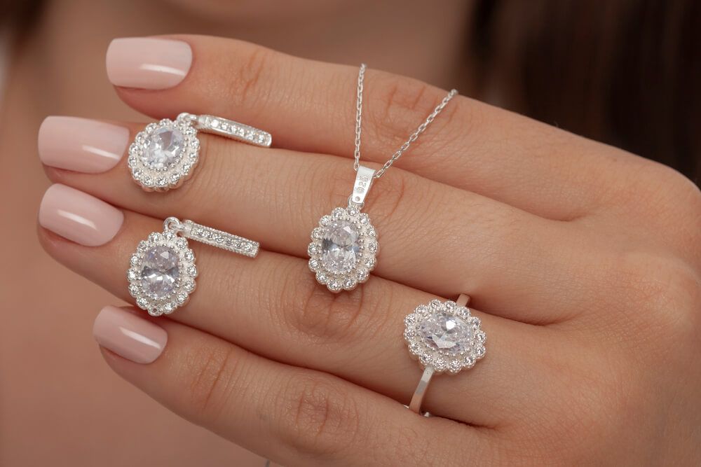 Woman's hand displaying a silver jewelry set featuring oval halo earrings, necklace, and ring with clear stones.