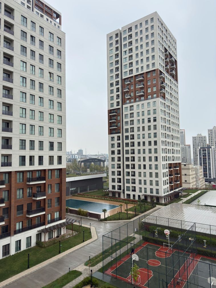 Two modern high-rise buildings, white and brown, with a sports court and pool in a city setting.