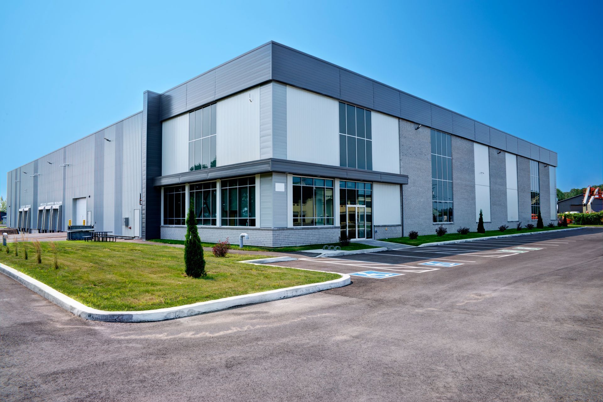 Modern gray and white industrial building with large windows and a paved driveway under a blue sky.