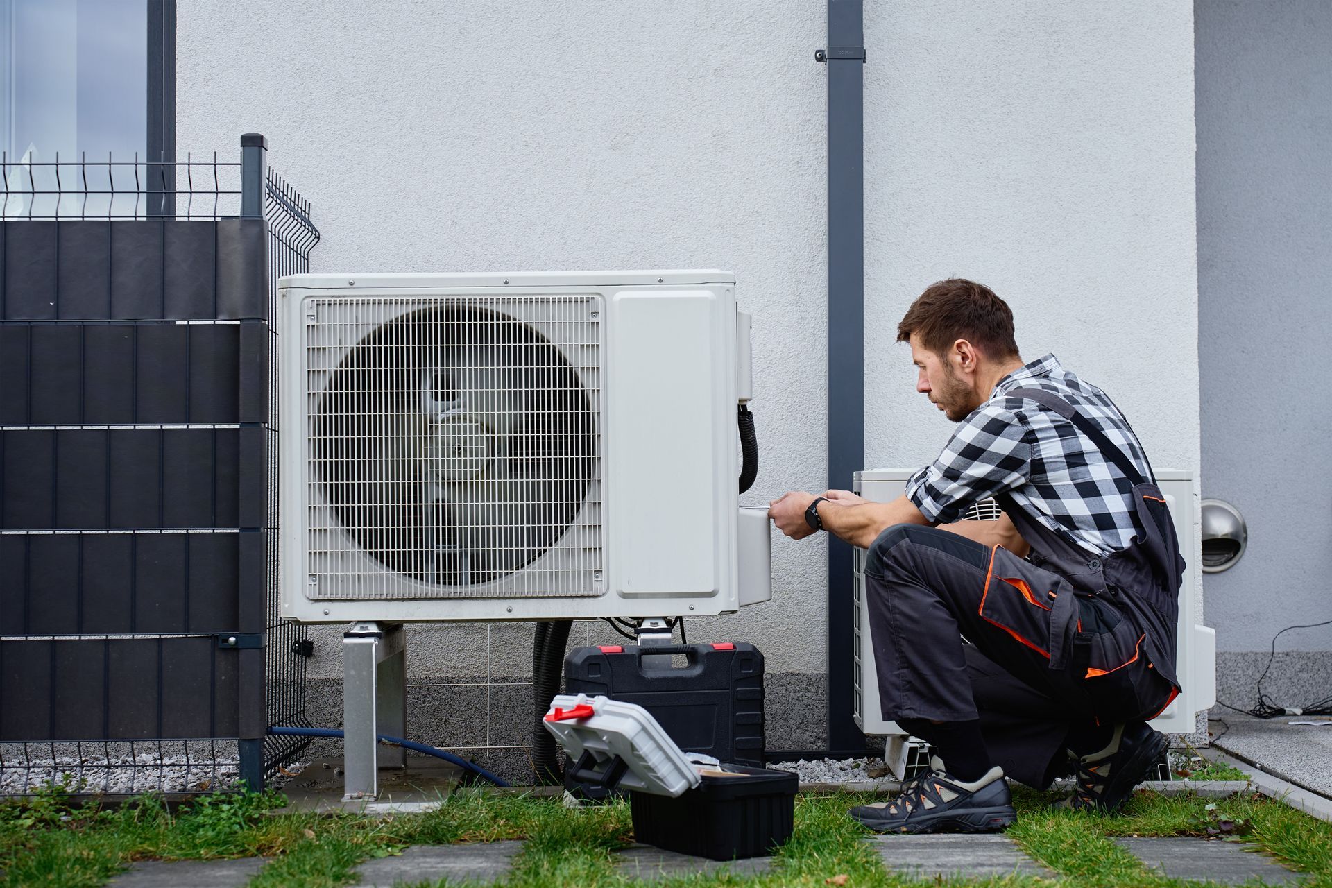 Technician crouching beside an outdoor air conditioner unit, inspecting it with tools near a building wall