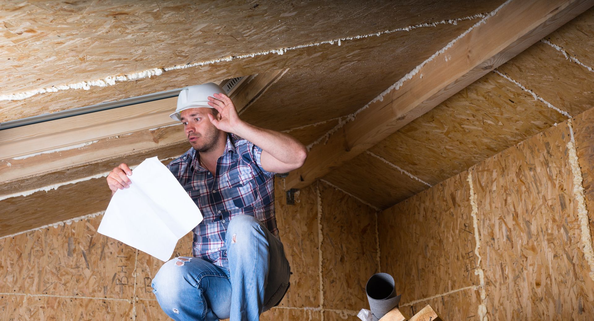 Worker in a hard hat inspecting insulation with exposed beams and papers in hand