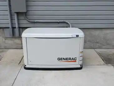 Outdoor beige generator beside a house, next to a white railing on gravel and grass