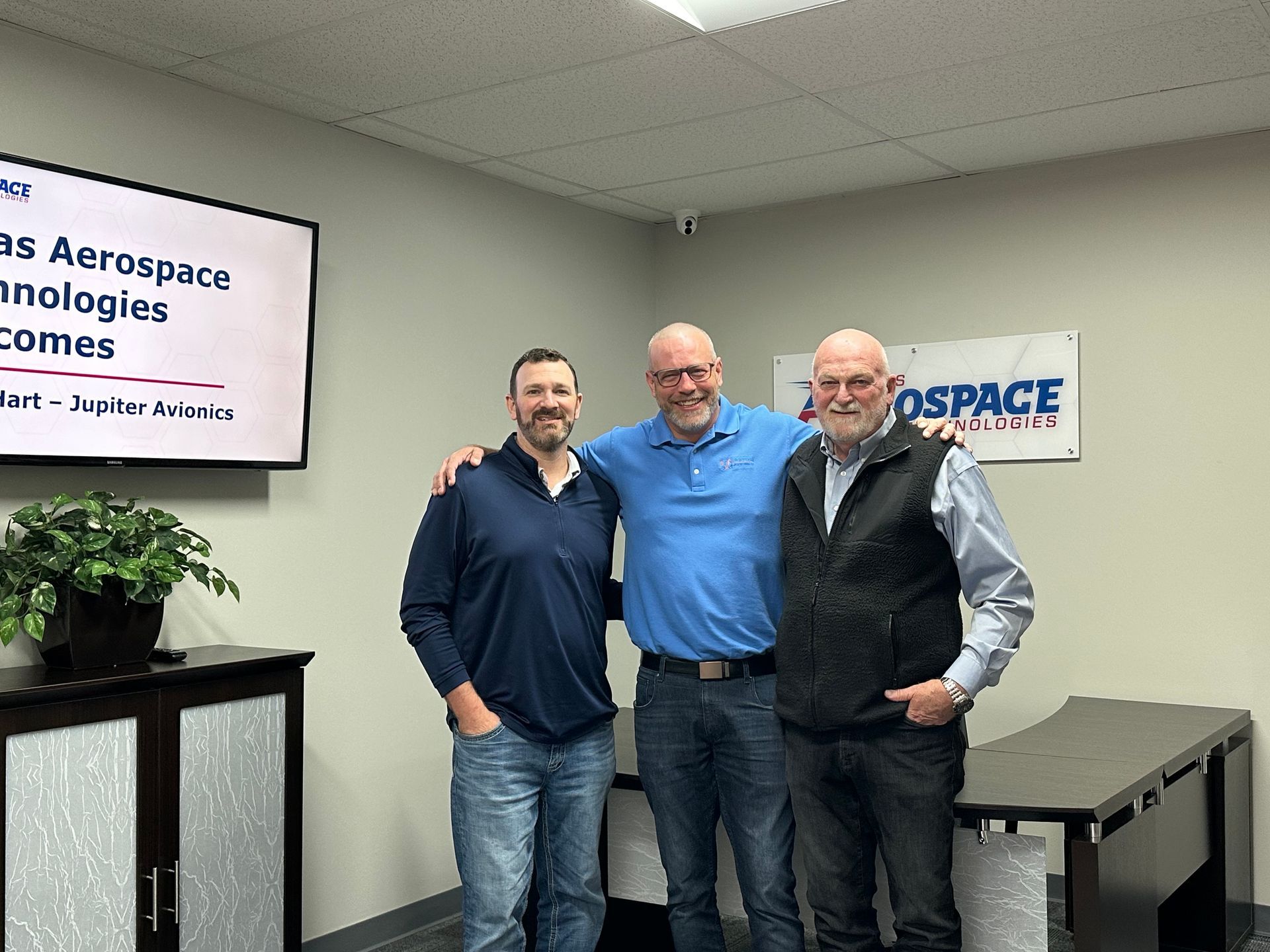 Three men stand smiling in an office, with aerospace signs and a table behind them.