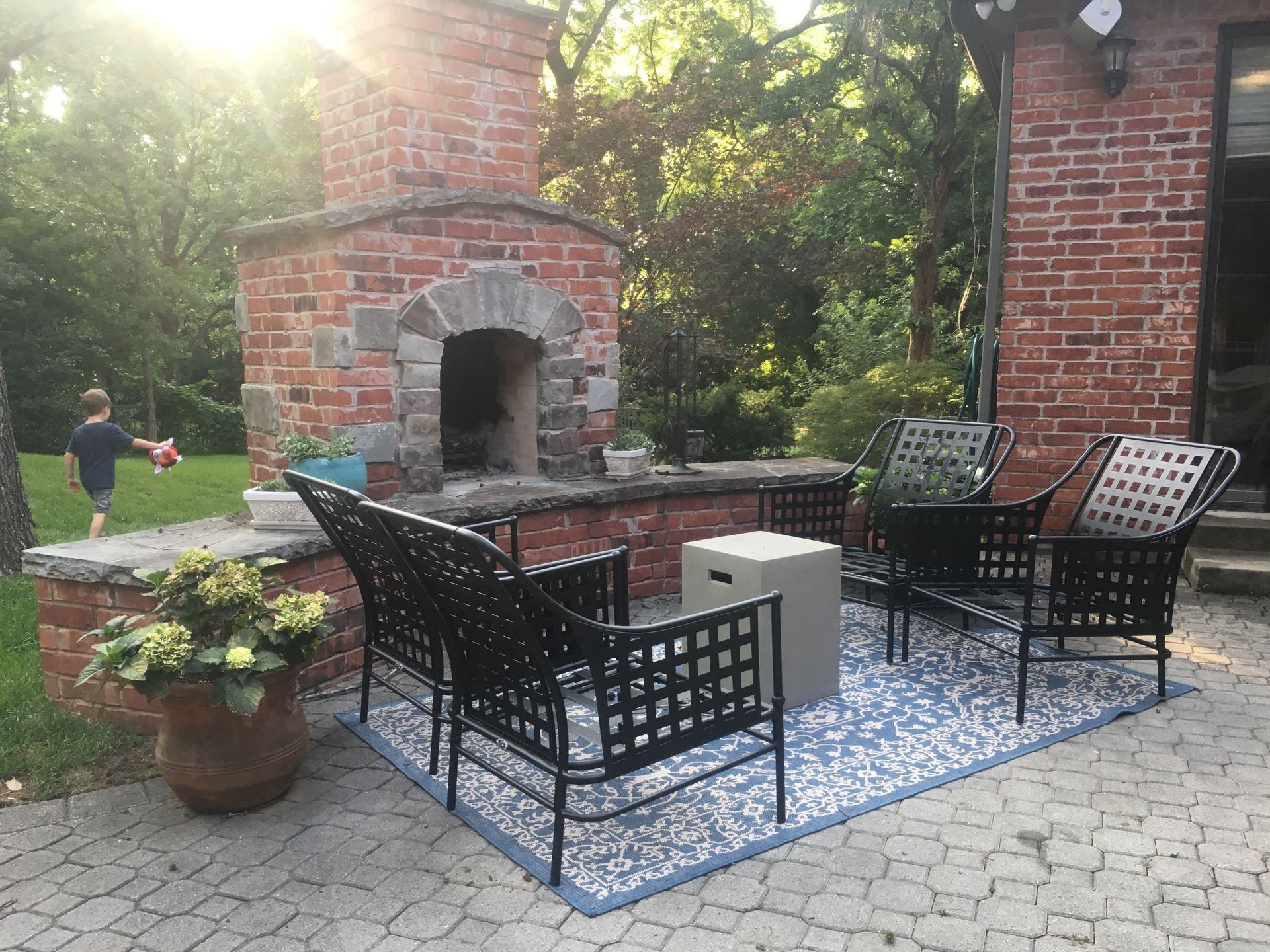 A brick fireplace is surrounded by chairs and a rug on a patio.