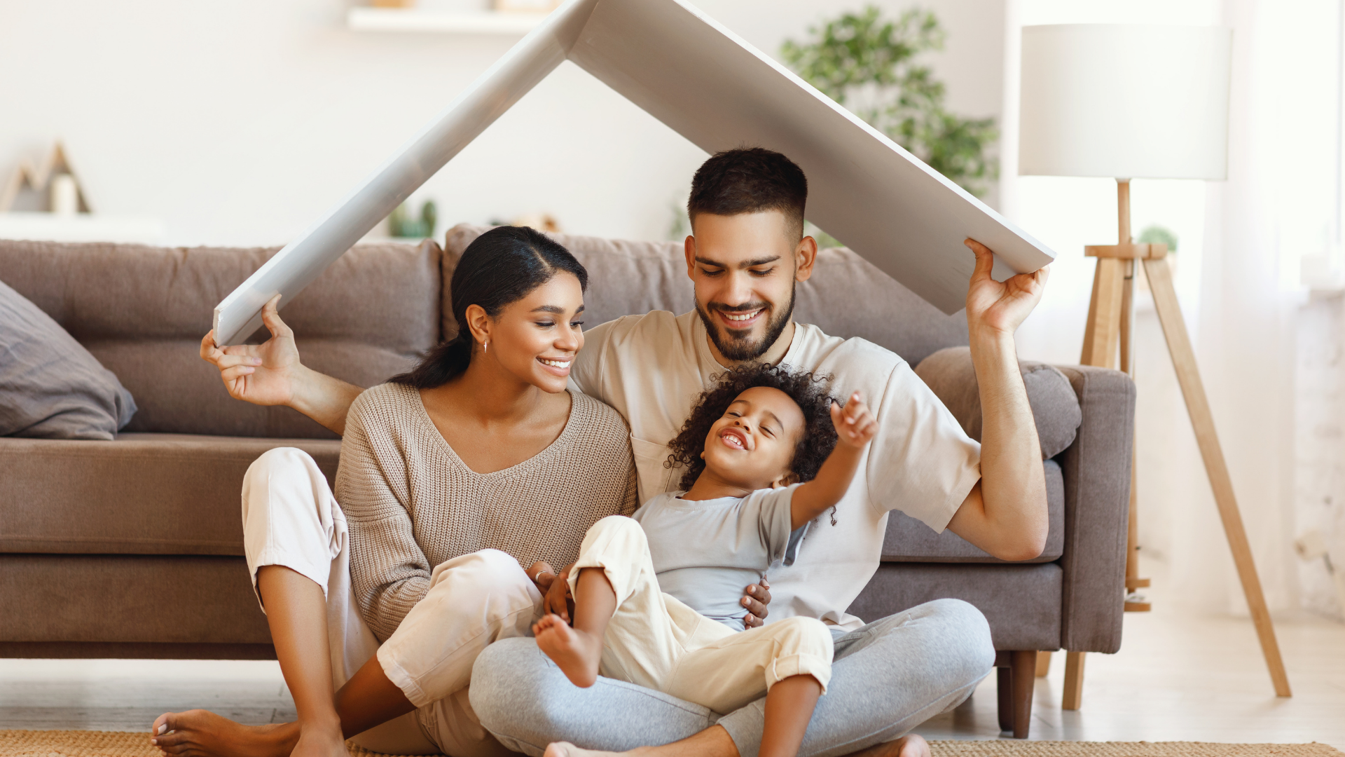 Family sitting on the floor under a tent made from a sofa and blanket in a bright living room.
