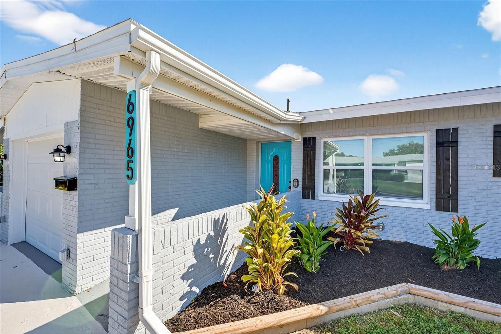 A light gray brick house exterior with a teal front door and house number 6965, featuring landscaping and a garage.