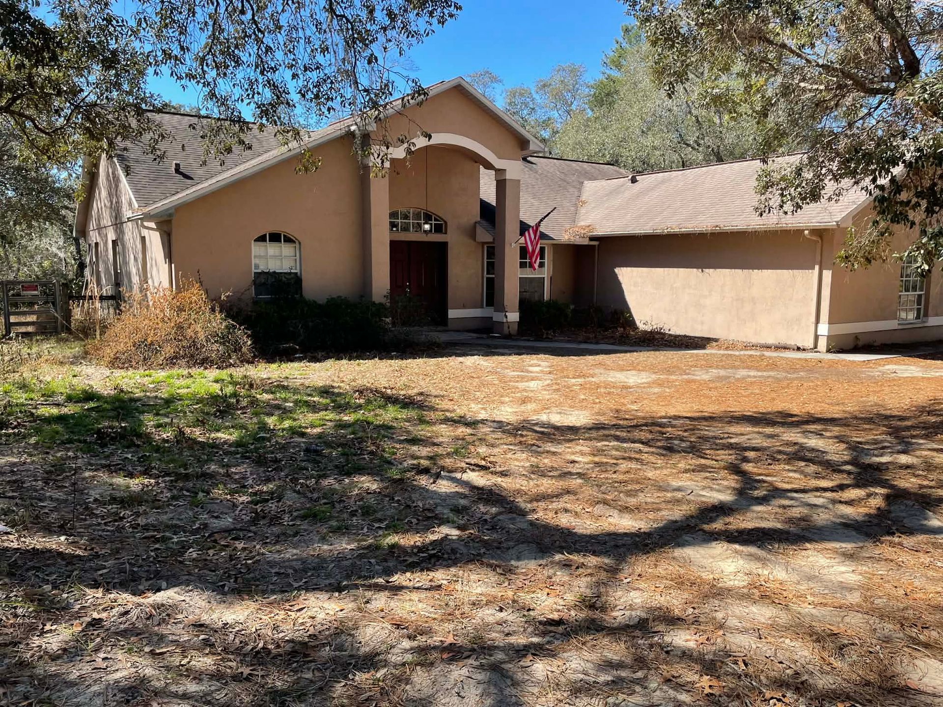 Tan stucco house with brown roof and bare yard, trees overhead.
