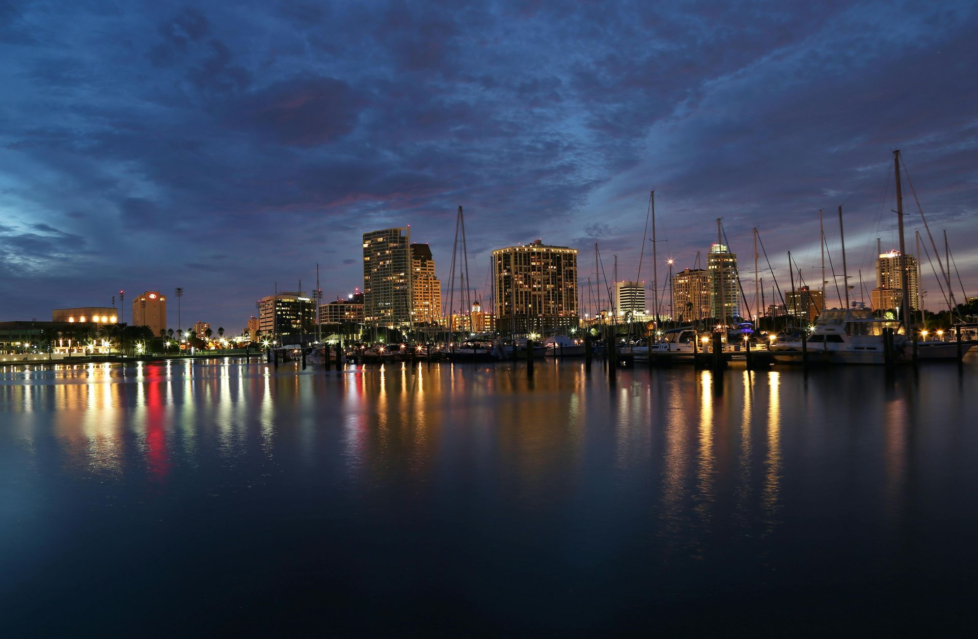 City skyline at dusk reflected in calm harbor waters, with boat masts silhouetted against a deep blue, cloudy sky.