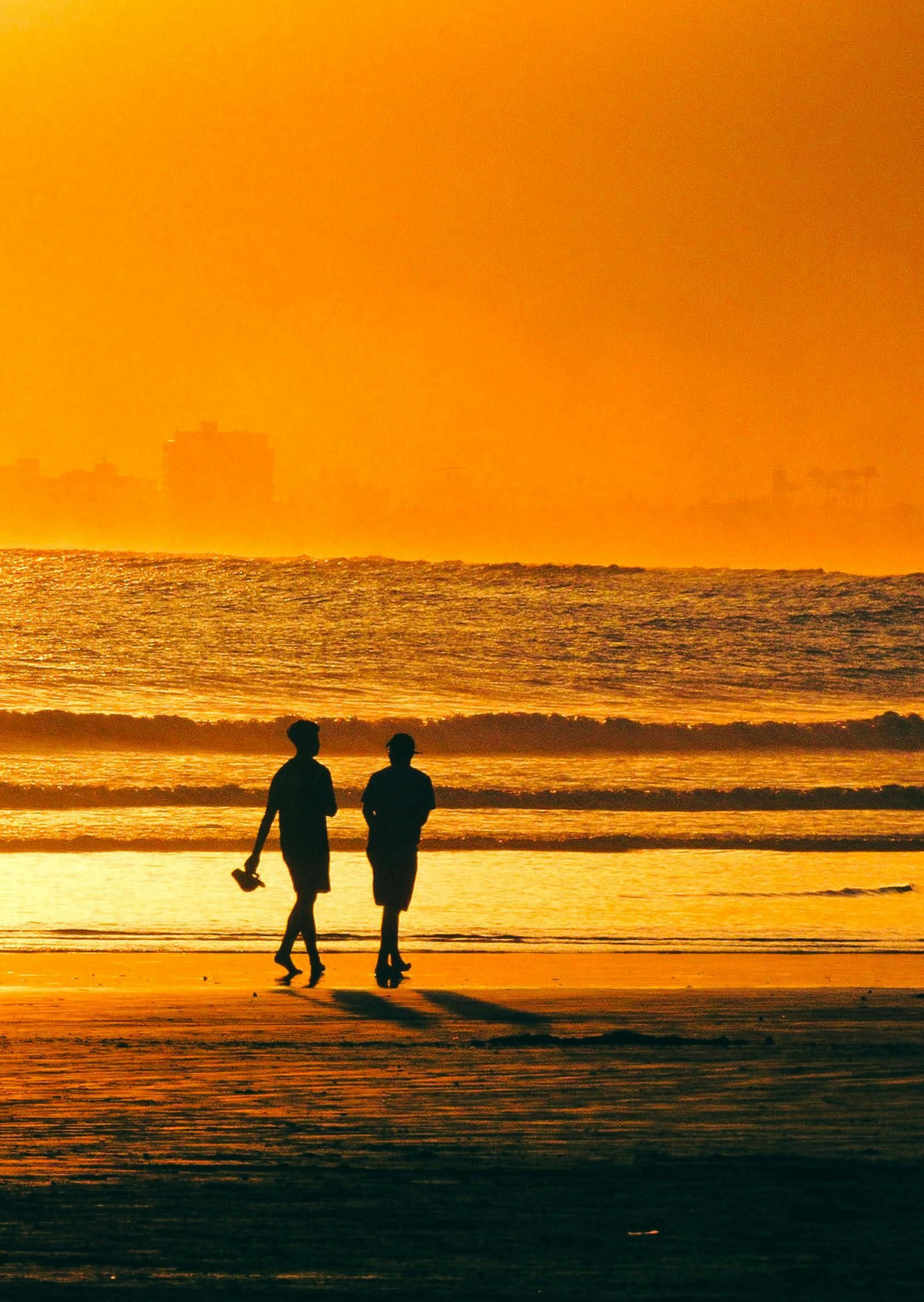 Silhouetted figures walk on a beach at sunset; orange sky, waves, distant buildings.