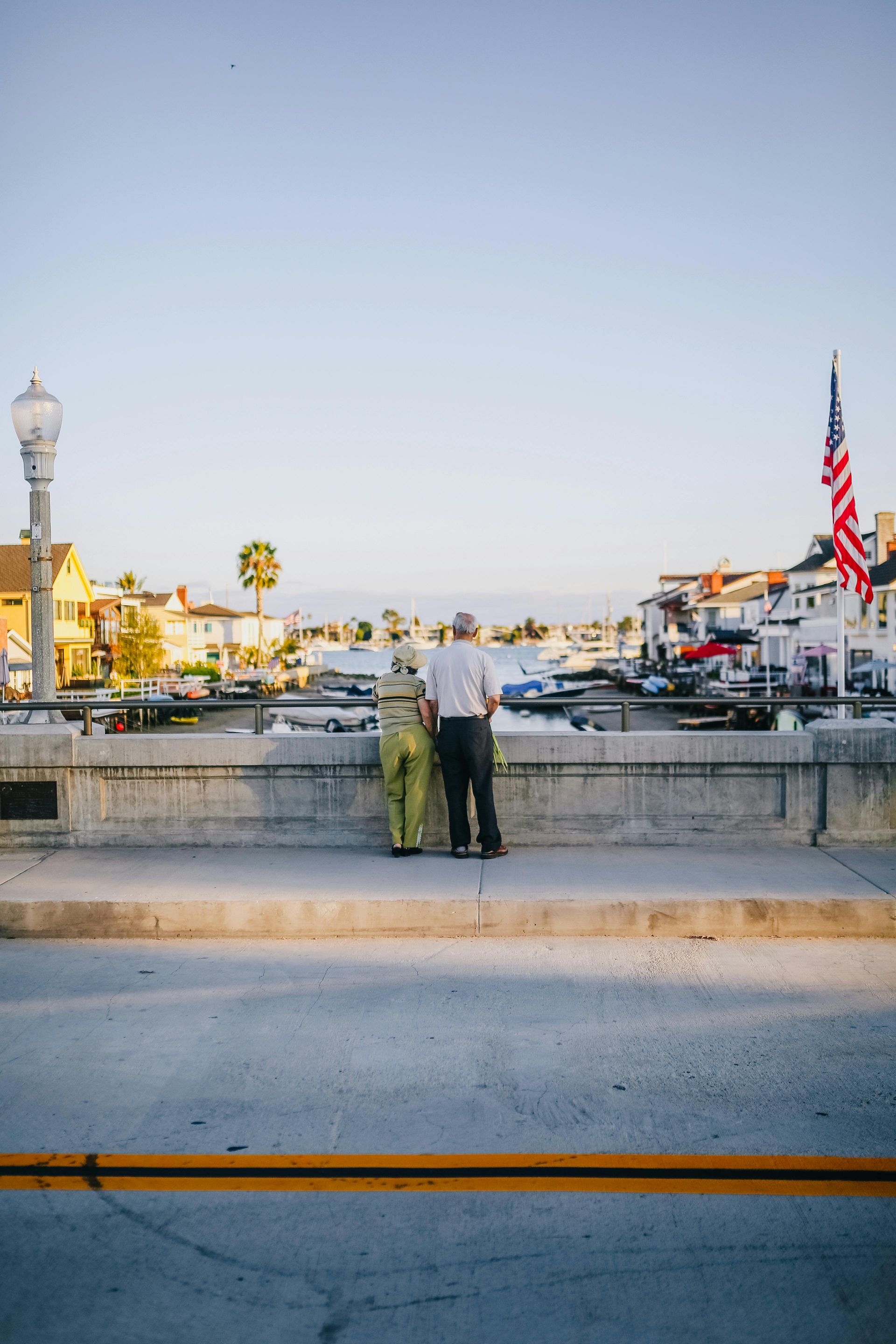 Couple standing on a bridge, overlooking a harbor with boats. American flag in the background.