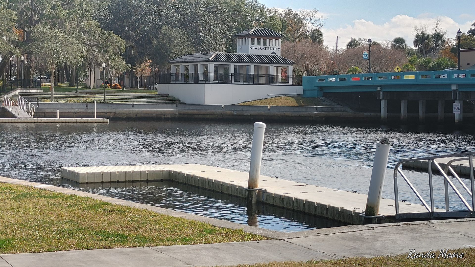 Dock on a waterway with a white building, trees, and a blue bridge in the background.