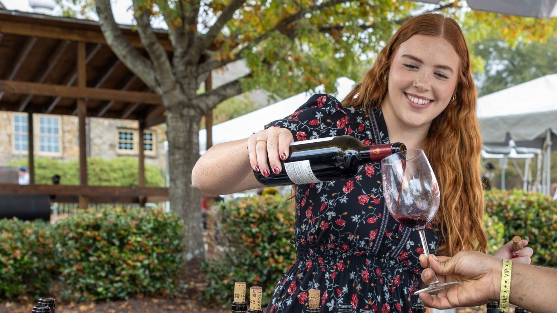 Woman pouring red wine into a glass outdoors. She smiles. The background includes a gazebo and greenery.
